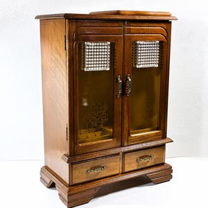 May include: A wooden jewelry armoire with a warm brown finish. It features two glass doors with decorative floral etching and small square accents. Two drawers are located at the bottom. The armoire has brass-coloured hardware and a rectangular shape.