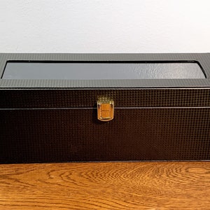 May include: A black rectangular watch box with a clear glass top and a gold latch. The box has a textured carbon fiber pattern.