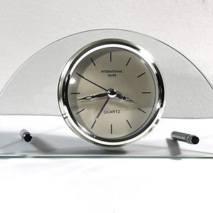 May include: A silver-toned analog clock with a glass face and a glass arch-shaped stand. The clock face has black hands and black Roman numeral hour markers. The clock face also has the text "INTERNATIONAL SILVER QUARTZ".