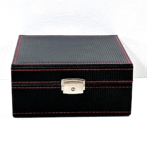 May include: A rectangular black storage box with a carbon fiber-like texture and contrasting red stitching along the edges. The box features a silver-toned metal latch centered on the front panel to secure the lid, providing a clean, structured look.