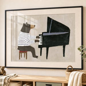 May include: A framed wall art print featuring a brown bear wearing a white and grey striped shirt, sitting on a small stool and playing a black grand piano. The illustration is set against a neutral beige background with a thin black frame.