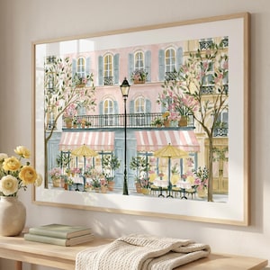 May include: A framed wall art print depicting a street scene with a pink building, a black street lamp, and a cafe with pink and white striped awnings. The scene includes yellow umbrellas, outdoor tables, and potted flowers in shades of pink.
