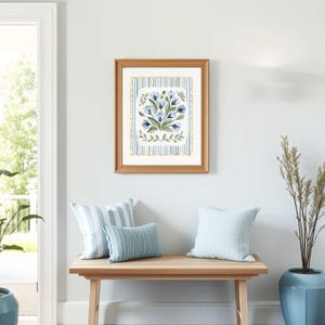 Puede incluir: Una impresi&oacute;n floral enmarcada con flores azules y verdes cuelga sobre un banco de madera con tres cojines. Los cojines son de rayas azules y blancas. Una planta alta en una maceta azul se encuentra a la derecha del banco. La habitaci&oacute;n tiene una pared azul claro y una puerta blanca.
