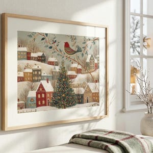May include: A framed horizontal wall art print depicting a winter village scene with snow-covered houses, a central decorated Christmas tree, and a red bird perched on a branch. The artwork features a muted colour palette with soft tones of white, grey, and red.