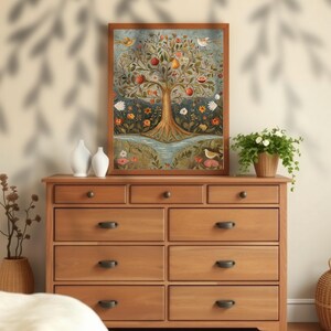 May include: A framed painting depicting a stylised tree laden with colourful fruits, flowers and birds. The tree is set against a blue background with a river flowing beneath it. The painting is displayed on a wooden dresser with other decorative items.