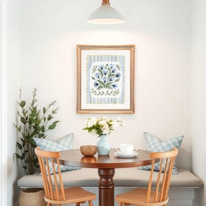 Puede incluir: Un rinc&oacute;n de comedor acogedor con una mesa redonda de madera y dos sillas de madera. Una impresi&oacute;n floral enmarcada cuelga de la pared, y un jarr&oacute;n con flores blancas est&aacute; sobre la mesa. Dos cojines azules estampados descansan en un banco.