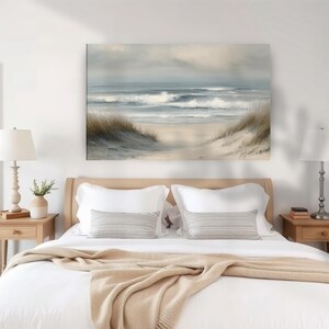 May include: A serene beach scene painting hangs above a bed with white linens and a tan blanket. The artwork depicts ocean waves, a sandy beach, and grassy dunes under a cloudy sky. Two bedside tables with lamps flank the bed.
