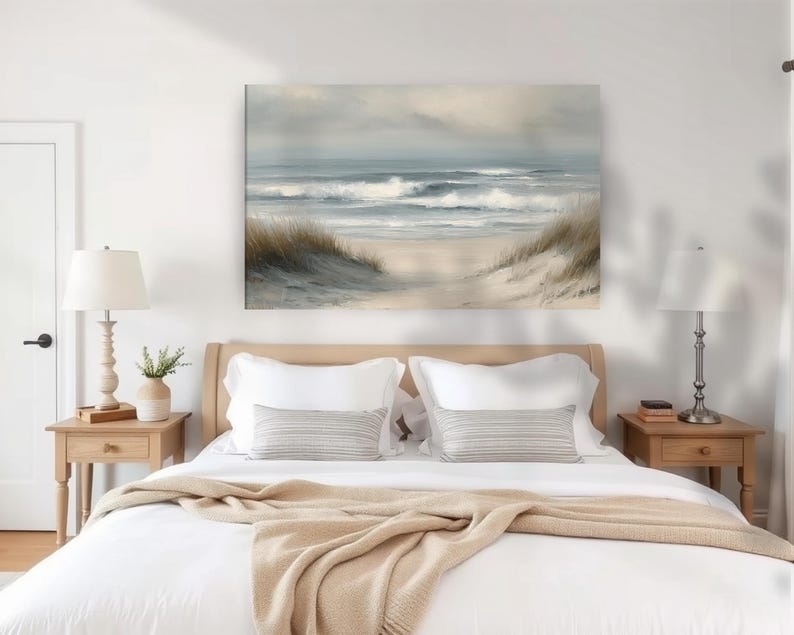 May include: A serene beach scene painting hangs above a bed with white linens and a tan blanket. The artwork depicts ocean waves, a sandy beach, and grassy dunes under a cloudy sky. Two bedside tables with lamps flank the bed.