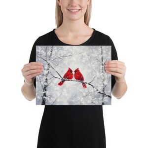 May include: A print featuring two vibrant red cardinals perched on a snow-covered branch. The artwork depicts a winter scene with a soft, snowy background and bare tree branches. The birds are the focal point, adding a pop of color.