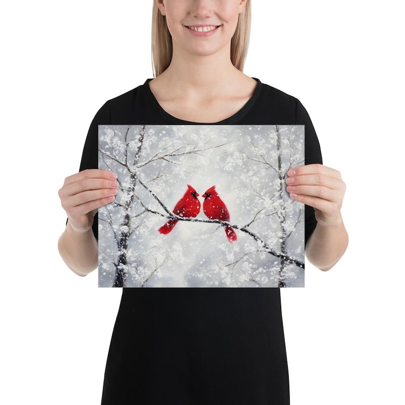 May include: A print featuring two vibrant red cardinals perched on a snow-covered branch. The artwork depicts a winter scene with a soft, snowy background and bare tree branches. The birds are the focal point, adding a pop of color.