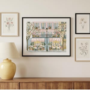 May include: A gallery wall arrangement featuring a large central framed print of a Parisian cafe scene with pink building facades, striped awnings, and yellow umbrellas, flanked by three smaller framed botanical sketches of flowers.