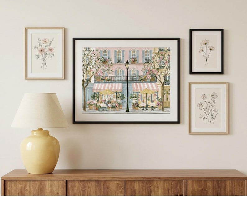 May include: A gallery wall arrangement featuring a large central framed print of a Parisian cafe scene with pink building facades, striped awnings, and yellow umbrellas, flanked by three smaller framed botanical sketches of flowers.