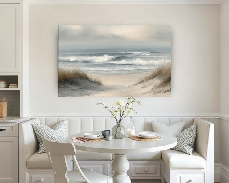 May include: A coastal landscape painting depicts a serene beach scene. The artwork features a sandy shore, rolling waves, and a cloudy sky. The painting is displayed above a white round table with a built-in bench seating area.