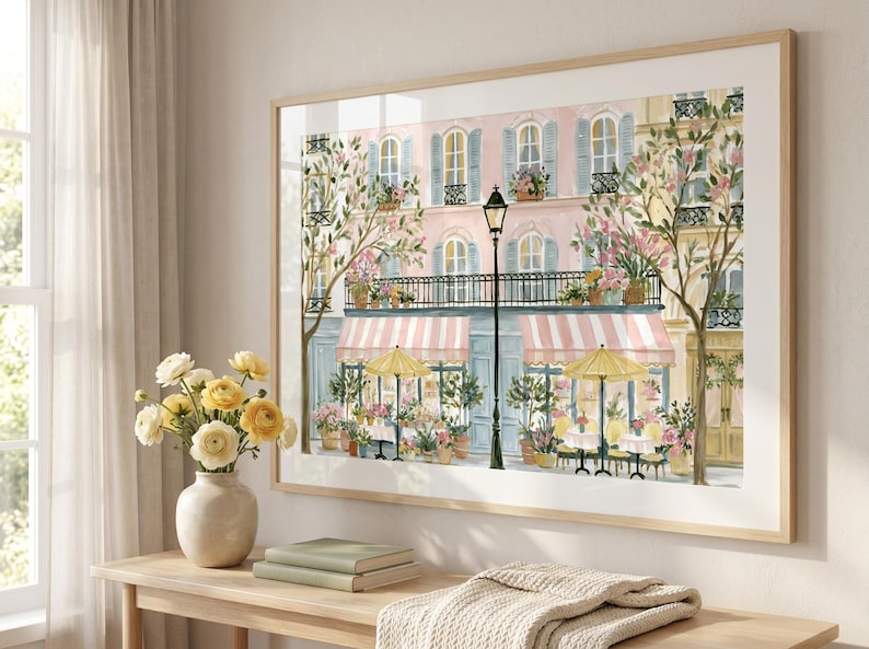 May include: A framed wall art print depicting a street scene with a pink building, a black street lamp, and a cafe with pink and white striped awnings. The scene includes yellow umbrellas, outdoor tables, and potted flowers in shades of pink.