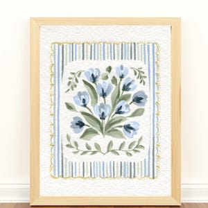Puede incluir: Una pintura de acuarela enmarcada con un ramo de flores azules con hojas verdes. La obra de arte est&aacute; sobre un fondo de rayas verticales azules y blancas, enmarcada por un borde dorado festoneado. El marco es de madera clara.
