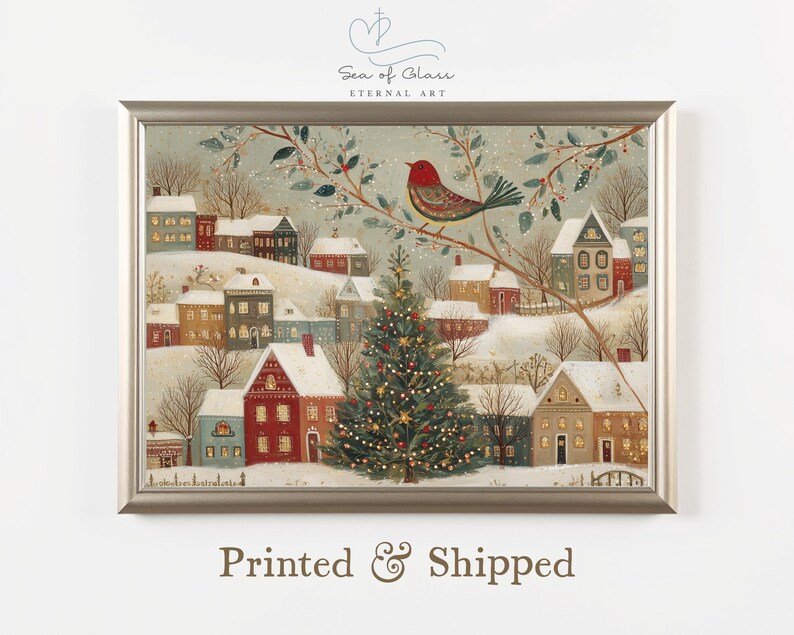 May include: A framed horizontal art print depicting a snowy winter village scene at twilight. A large, lit Christmas tree stands in the centre, surrounded by houses with glowing windows. A red bird sits on a branch in the foreground.