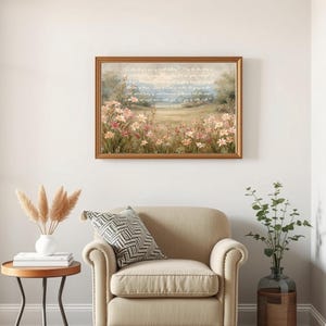 May include: A framed landscape painting with a field of pink and white flowers, a blue sky, and text. The painting hangs above a beige armchair with a patterned pillow. A small side table with a vase of dried flowers and books sits nearby.