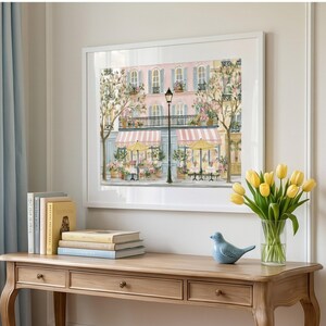 May include: A framed wall art print depicting a Parisian street scene with a pink building, striped awnings, yellow umbrellas, and a black street lamp. A wooden console table holds a stack of books, a blue ceramic bird, and a vase of yellow tulips.