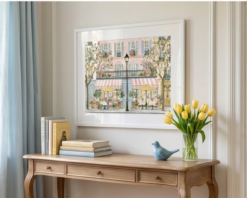 May include: A framed wall art print depicting a Parisian street scene with a pink building, striped awnings, yellow umbrellas, and a black street lamp. A wooden console table holds a stack of books, a blue ceramic bird, and a vase of yellow tulips.