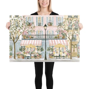 May include: A rectangular art print depicting a Parisian-style street scene with a pink building, a black lamppost, two yellow umbrellas, and outdoor cafe tables. The storefront features striped pink and white awnings and potted flowers.