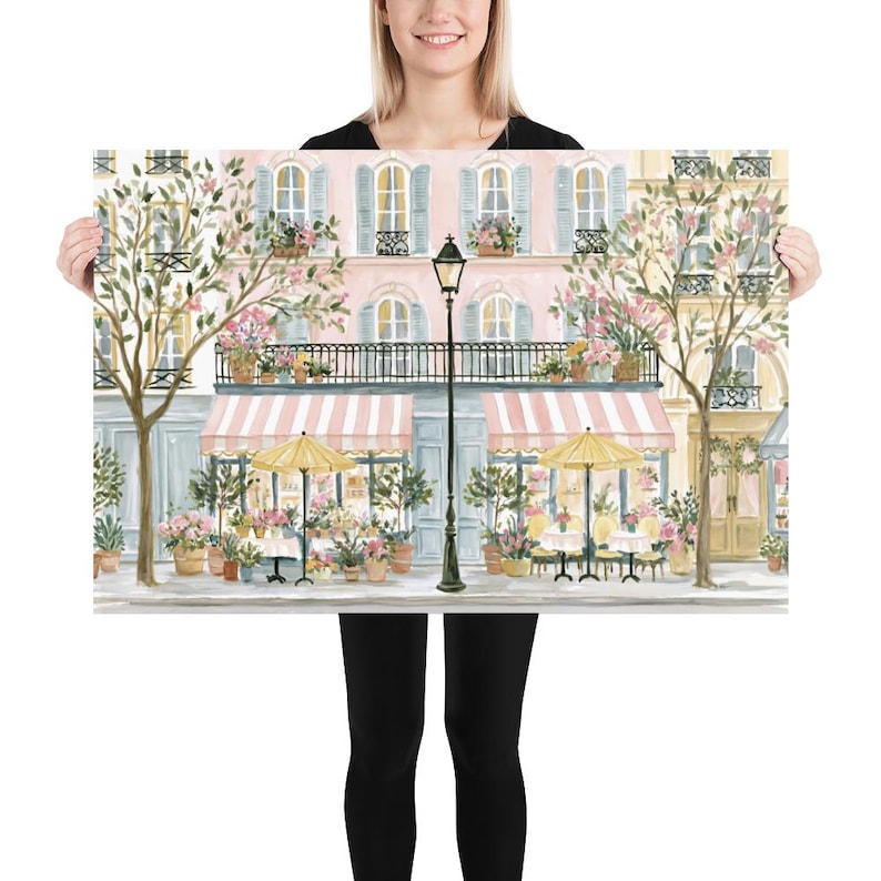 May include: A rectangular art print depicting a Parisian-style street scene with a pink building, a black lamppost, two yellow umbrellas, and outdoor cafe tables. The storefront features striped pink and white awnings and potted flowers.