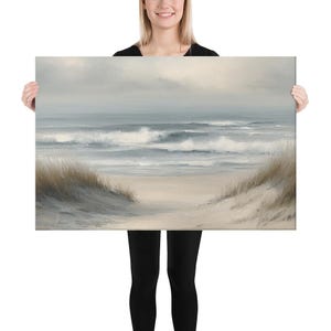 May include: A canvas print of a serene beach scene. The artwork depicts a sandy beach with dunes and tall grass, leading to a calm ocean under a cloudy sky. The color palette is soft, with muted blues, grays, and beige tones.