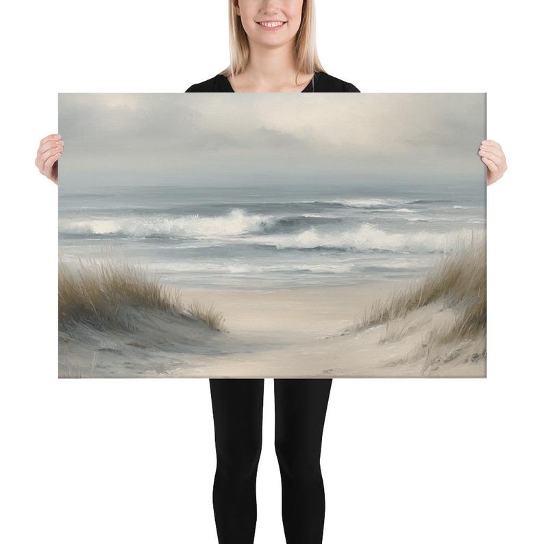 May include: A canvas print of a serene beach scene. The artwork depicts a sandy beach with dunes and tall grass, leading to a calm ocean under a cloudy sky. The color palette is soft, with muted blues, grays, and beige tones.