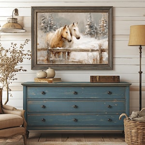 May include: A framed painting depicts two horses in a snowy landscape, with a wooden fence. The painting hangs above a distressed blue dresser with three drawers. A floor lamp with a tan shade and a wicker basket are also in the room.