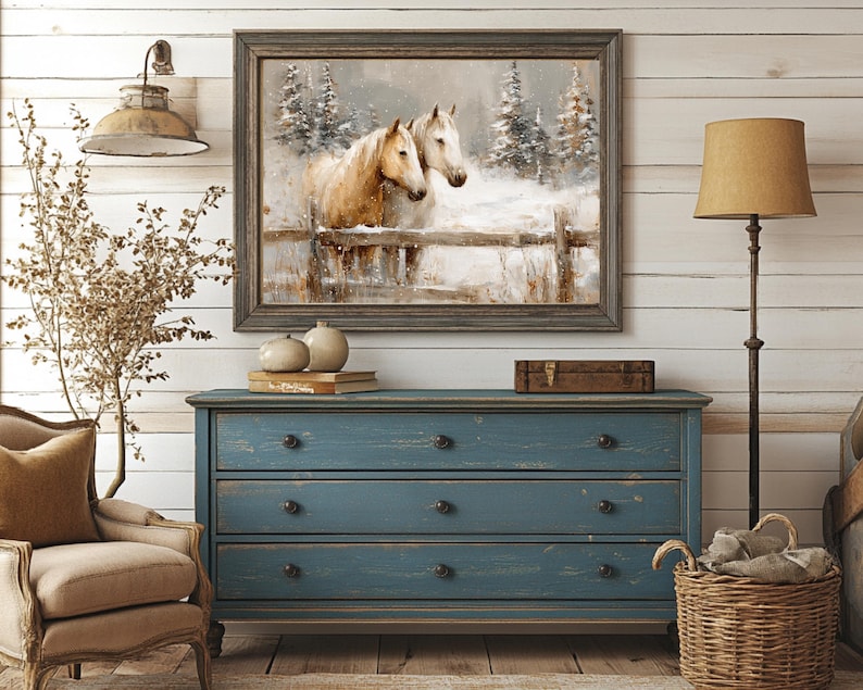 May include: A framed painting depicts two horses in a snowy landscape, with a wooden fence. The painting hangs above a distressed blue dresser with three drawers. A floor lamp with a tan shade and a wicker basket are also in the room.