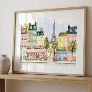 May include: Watercolor painting of a Parisian street scene with colorful buildings, a cafe, and the Eiffel Tower. The artwork is framed in a light wood frame and displayed on a wooden shelf. A decorative vase sits on the shelf.