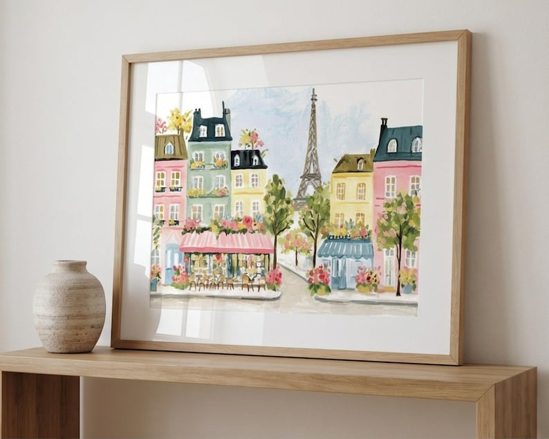 May include: Watercolor painting of a Parisian street scene with colorful buildings, a cafe, and the Eiffel Tower. The artwork is framed in a light wood frame and displayed on a wooden shelf. A decorative vase sits on the shelf.