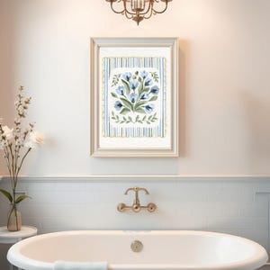 Puede incluir: Una impresi&oacute;n floral enmarcada con flores azules y hojas verdes. La obra de arte est&aacute; sobre un fondo a rayas con un borde festoneado. La impresi&oacute;n cuelga sobre una ba&ntilde;era blanca con accesorios dorados.