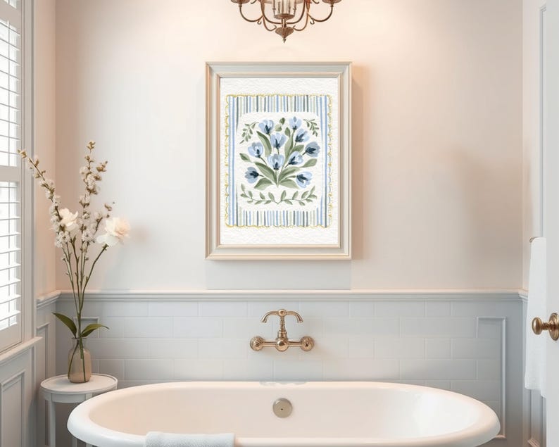 Puede incluir: Una impresi&oacute;n floral enmarcada con flores azules y hojas verdes. La obra de arte est&aacute; sobre un fondo a rayas con un borde festoneado. La impresi&oacute;n cuelga sobre una ba&ntilde;era blanca con accesorios dorados.