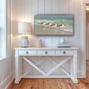 May include: A rectangular canvas art piece depicts four sandpipers walking along a sandy beach with gentle waves in the background. The artwork is displayed above a white console table with three drawers and a decorative X-shaped base.