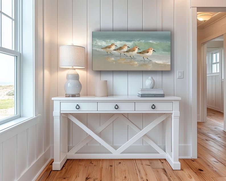 May include: A rectangular canvas art piece depicts four sandpipers walking along a sandy beach with gentle waves in the background. The artwork is displayed above a white console table with three drawers and a decorative X-shaped base.