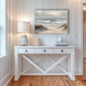 May include: A coastal-themed interior featuring a white console table with three drawers, a decorative lamp, and a framed painting of a beach scene. The painting depicts a sandy path leading to the ocean under a cloudy sky. The room has white paneling and hardwood floors.