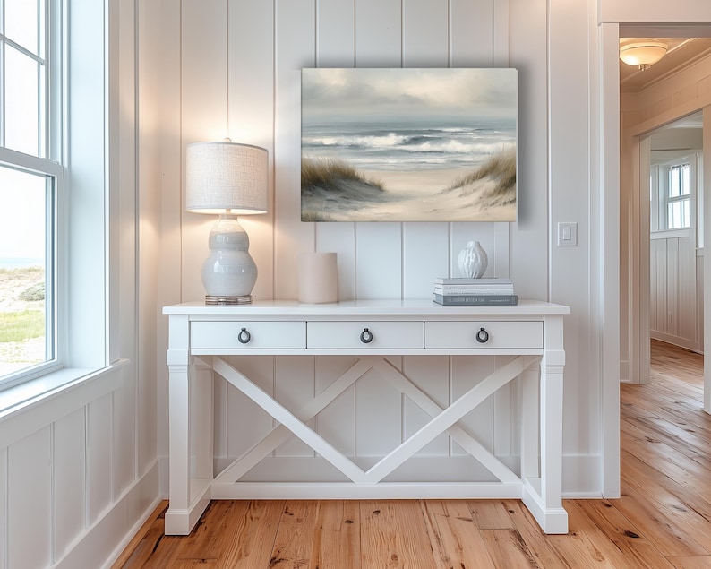 May include: A coastal-themed interior featuring a white console table with three drawers, a decorative lamp, and a framed painting of a beach scene. The painting depicts a sandy path leading to the ocean under a cloudy sky. The room has white paneling and hardwood floors.