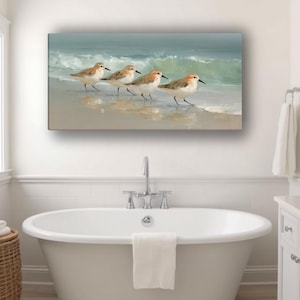 May include: A rectangular wall art print depicts four sandpipers walking along a sandy beach with gentle waves. The artwork features soft, muted colors of beige, blue, and brown, creating a serene coastal scene. The print is displayed above a white bathtub.
