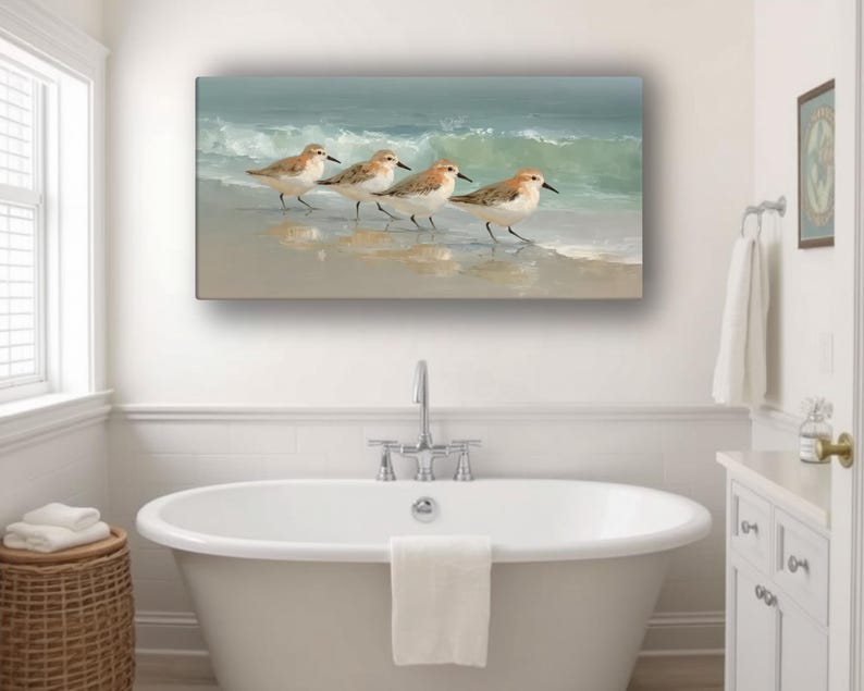 May include: A rectangular wall art print depicts four sandpipers walking along a sandy beach with gentle waves. The artwork features soft, muted colors of beige, blue, and brown, creating a serene coastal scene. The print is displayed above a white bathtub.