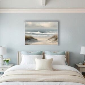 May include: A framed canvas art print depicts a serene beach scene. The artwork features a sandy path leading to the ocean, with gentle waves and a cloudy sky. The color palette includes soft blues, beige, and muted greens.