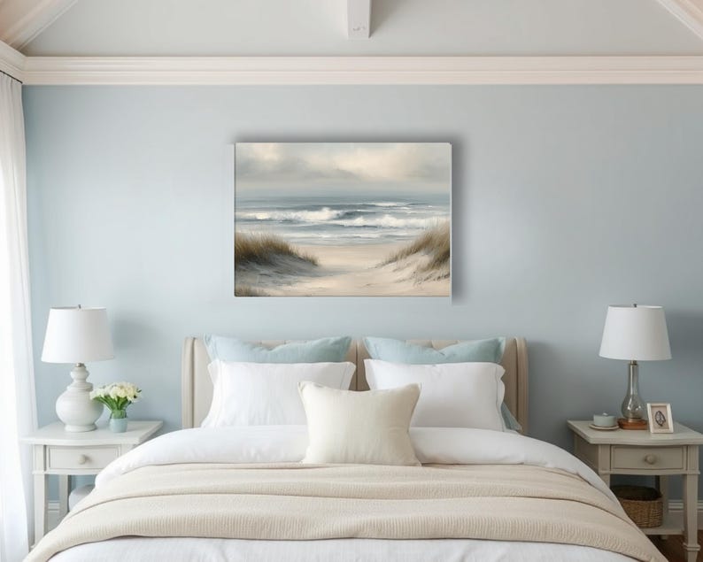 May include: A framed canvas art print depicts a serene beach scene. The artwork features a sandy path leading to the ocean, with gentle waves and a cloudy sky. The color palette includes soft blues, beige, and muted greens.