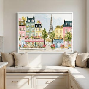 May include: Watercolor painting of a Parisian street scene with colorful buildings, a cafe, and the Eiffel Tower. The artwork is framed in white and displayed above a window seat with beige pillows.