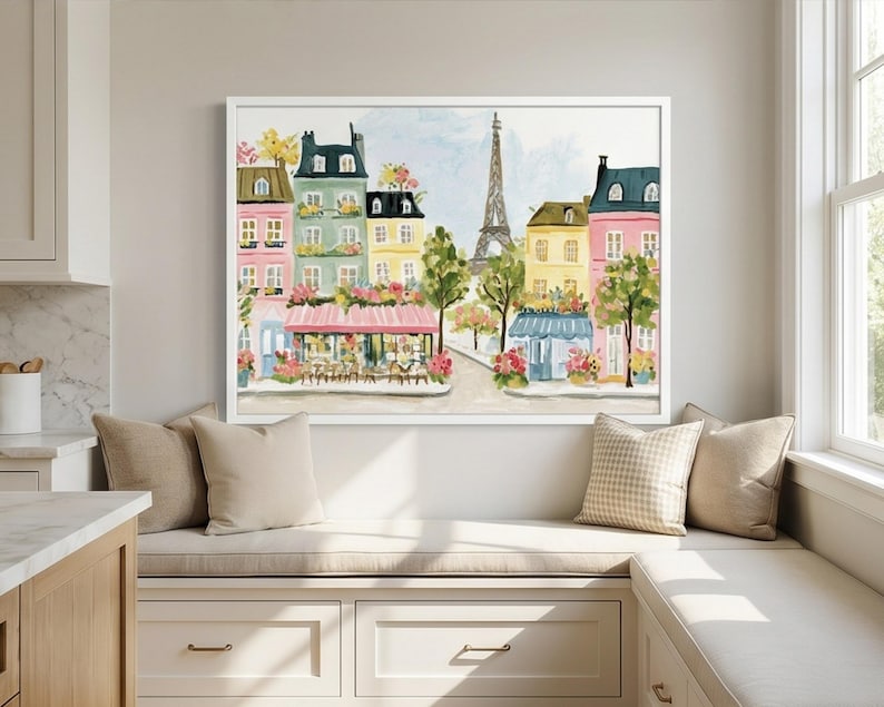 May include: Watercolor painting of a Parisian street scene with colorful buildings, a cafe, and the Eiffel Tower. The artwork is framed in white and displayed above a window seat with beige pillows.