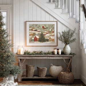 May include: A framed rectangular wall art print depicting a snowy winter village scene with colourful houses, a central evergreen tree, and a red bird perched on a branch. The artwork is displayed above a wooden console table with baskets.
