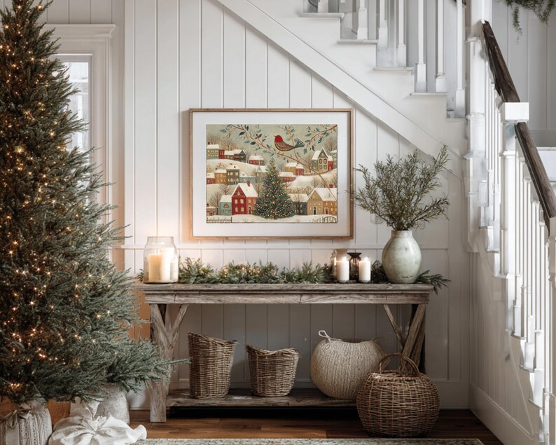 May include: A framed rectangular wall art print depicting a snowy winter village scene with colourful houses, a central evergreen tree, and a red bird perched on a branch. The artwork is displayed above a wooden console table with baskets.