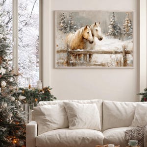 May include: A framed watercolor painting depicts two horses in a snowy landscape. The horses, with light brown coats, stand behind a wooden fence. Evergreen trees and falling snow complete the winter scene. The painting hangs above a white sofa, next to a decorated Christmas tree.