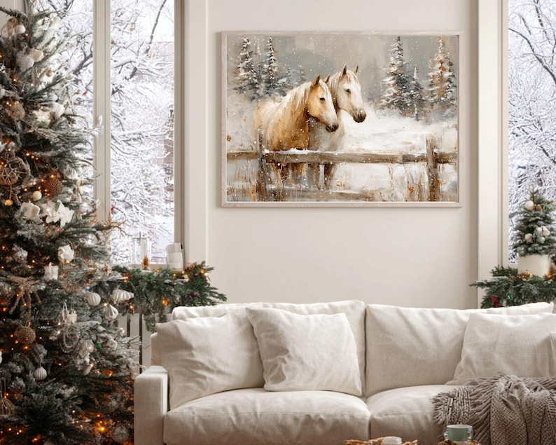 May include: A framed watercolor painting depicts two horses in a snowy landscape. The horses, with light brown coats, stand behind a wooden fence. Evergreen trees and falling snow complete the winter scene. The painting hangs above a white sofa, next to a decorated Christmas tree.