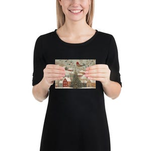 May include: A rectangular greeting card featuring a winter village scene with snow-covered houses, a central illuminated Christmas tree, and a small red bird perched on a branch above, all set against a soft, muted, and snowy landscape.