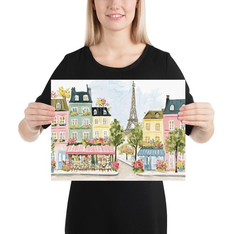 May include: Watercolor print of a Parisian street scene featuring colorful buildings, cafes, and the Eiffel Tower. The artwork showcases a charming street with pink, yellow, and green buildings adorned with flowers. A light blue sky provides the backdrop for this picturesque scene.