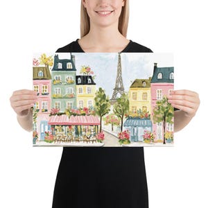 May include: A colorful print of a Parisian street scene featuring the Eiffel Tower. The artwork depicts buildings in shades of pink, green, and yellow, with flower boxes and a cafe. The print is held by a person.